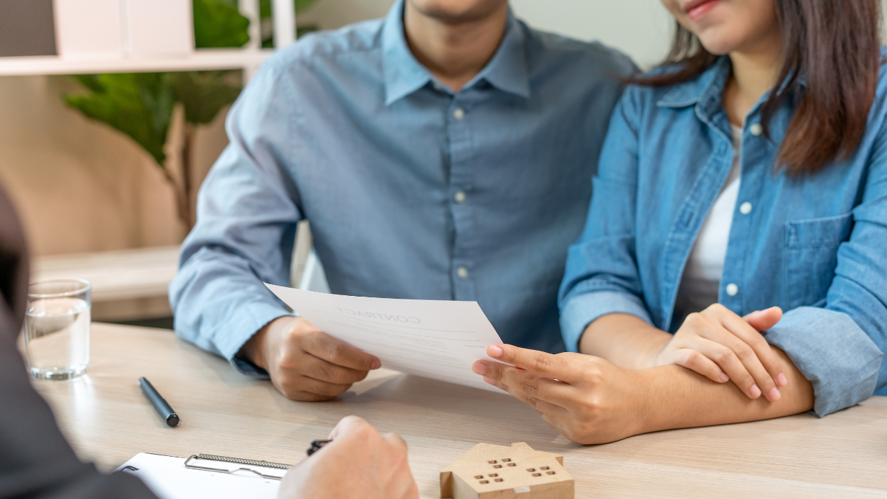 A couple in denim shirts is seated at a desk, holding and reviewing a document. A person in a black suit is in the foreground with a clipboard.