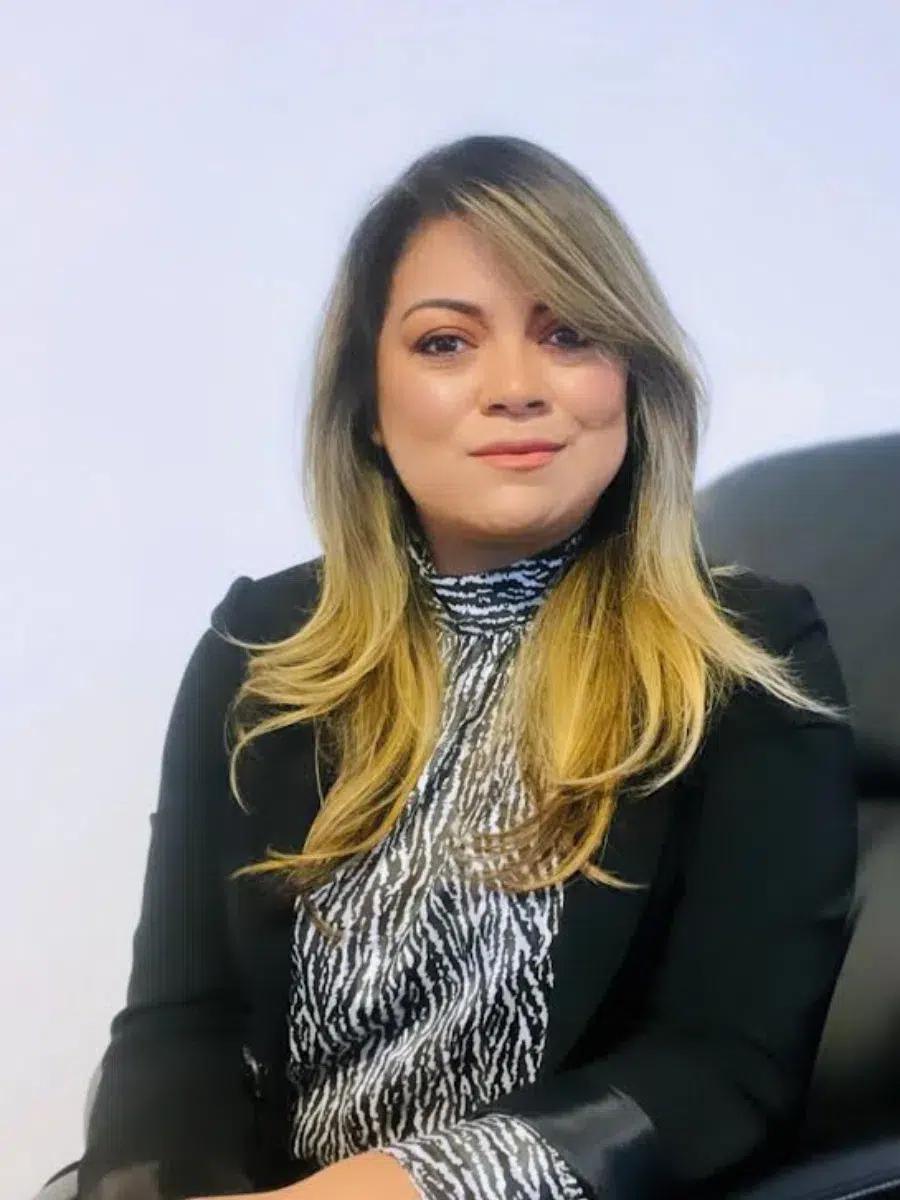 Person with long hair sits on a black chair, wearing a black blazer and patterned top, against a plain background.