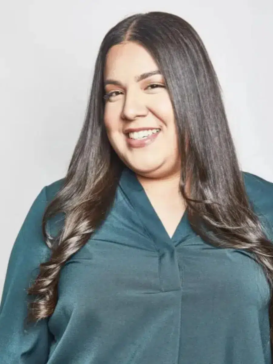 Smiling person with long dark hair wearing a teal top against a light background.