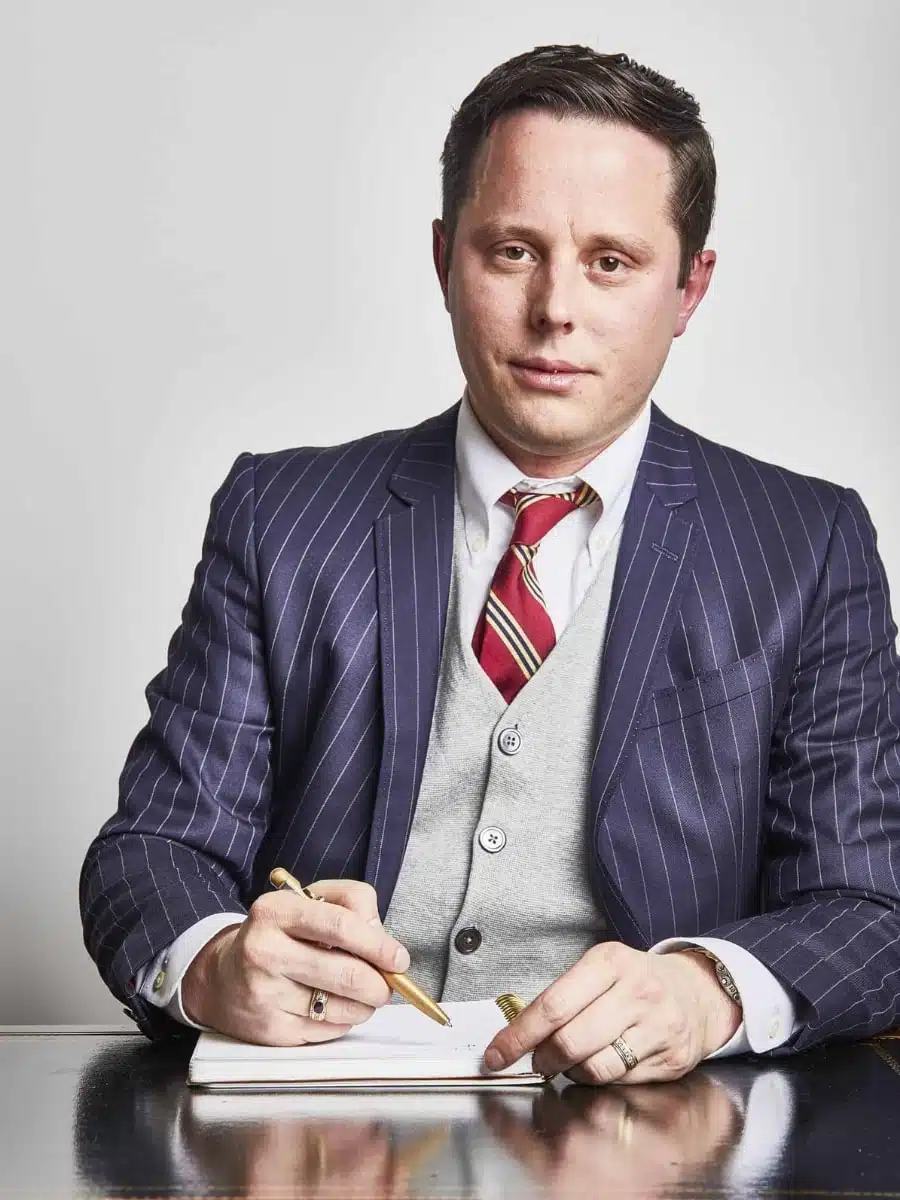 A man in a pinstripe suit and striped tie writes in a notebook with a pen, sitting at a desk.