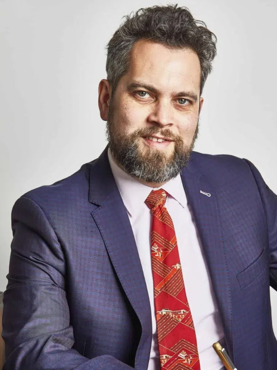 Man with a beard and mustache wearing a blue suit and red tie, holding a pen, sits against a plain background.