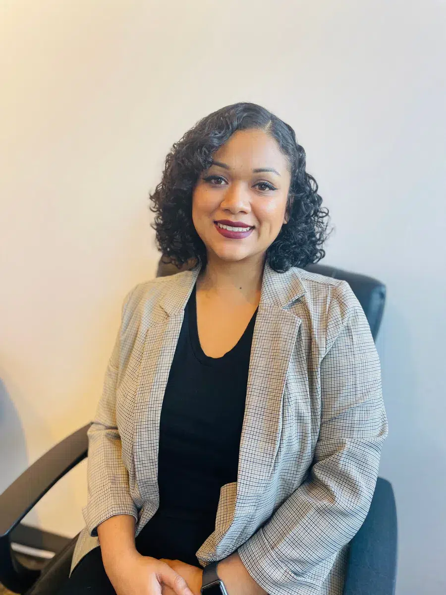 Person with curly hair in a blazer sits on a chair, smiling at the camera against a plain background.